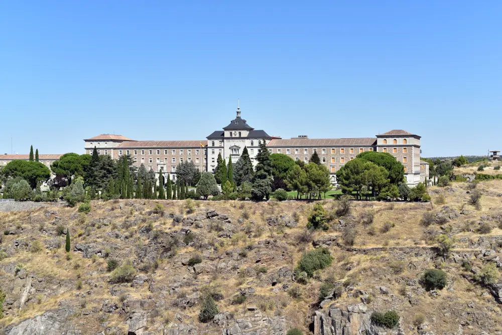 Imponente caserma e accademia militare in pietra su un'altura rocciosa circondata da alberi e vegetazione mediterranea.