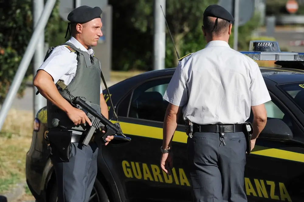 Allievo maresciallo della Guardia di Finanza in divisa con equipaggiamento tecnico e arma accanto a un'auto di pattuglia.