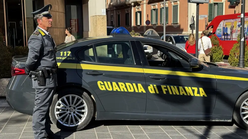 Ufficiale in uniforme della Guardia di Finanza con stivali e equipaggiamento tattico accanto a un'auto di servizio.
