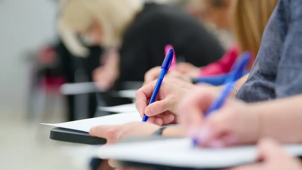 Primo piano di candidati che compilano i fogli durante la prova scritta di un concorso in aula d'esame.
