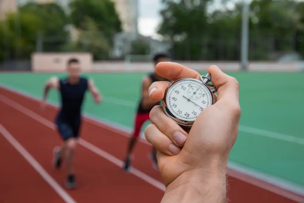 Mano con cronometro mentre dei candidati affrontano le prove fisiche di corsa su una pista di atletica.